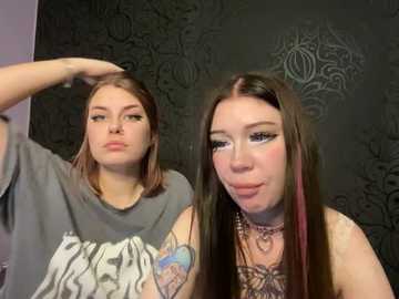Video of two young women with long, straight hair. One wears a grey graphic tee and has a tattooed arm. The other has dark hair and wears a black choker. They stand against a dark, patterned wallpaper background.