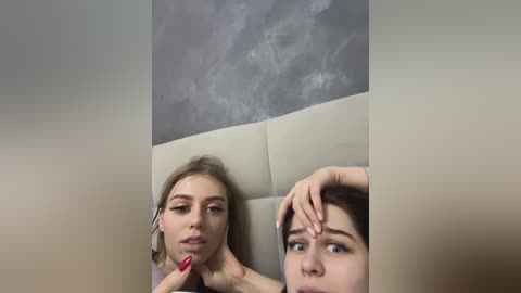 Media: A video shows two young women with pale skin and long hair, one with light blonde hair and another with dark hair, seated side by side on a cream-colored leather sofa. The background features a textured, gray wall.