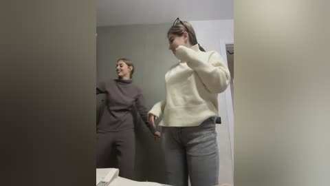 Media: Video of two women in a minimalist room. One in a beige sweater and jeans, the other in a grey sweater and pants, laughing.