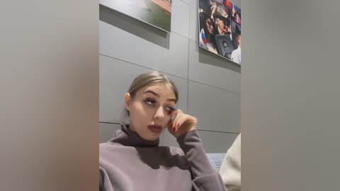Media: Video of a young woman with light brown hair, wearing a purple sweater, resting her chin on her hand in a museum with gray walls and framed photos.