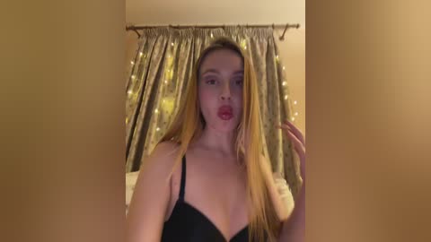 A video of a young, fair-skinned woman with long blonde hair, wearing black lingerie, in a bedroom with gold curtains and fairy lights.