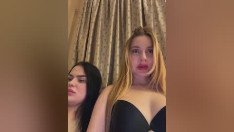 Video of two women in black strapless bras, one with long black hair, the other with blonde hair, standing in front of beige curtains in a dimly lit room.