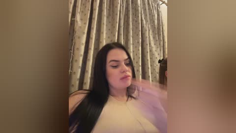 A video shows a fair-skinned woman with long black hair, wearing a beige top, sitting in front of patterned curtains. She looks relaxed, her eyes closed, and a hand near her face.
