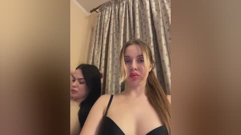 Video of two women in a bathroom. One has light skin, long blonde hair, and wears a black bra, while the other has dark hair and is partially visible in the background.