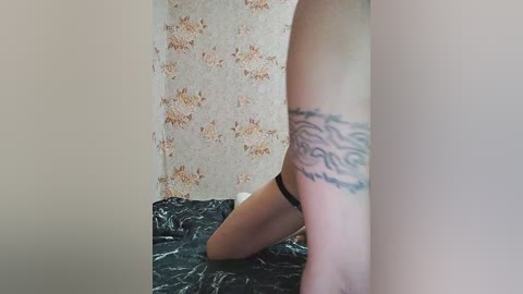 Video of a nude woman kneeling on a black marble-patterned bedspread, showing a tattoo on her thigh. She has light skin and is facing away from the camera. The background features floral wallpaper.