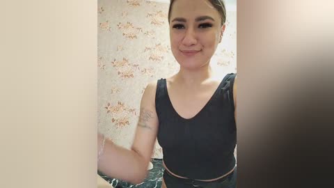 Video of a young woman with light skin and dark hair tied back, wearing a black sleeveless top and denim shorts. She smiles at the camera, standing in a room with floral wallpaper and a blurred brown door in the background.