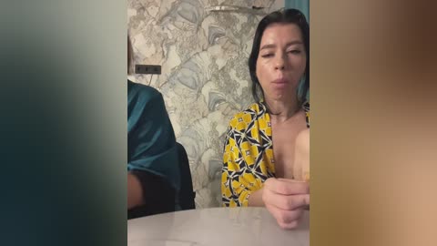 Media: A video captures a woman with dark hair, wearing a yellow and black patterned top, blowing a bubble, in a bathroom with floral wallpaper.