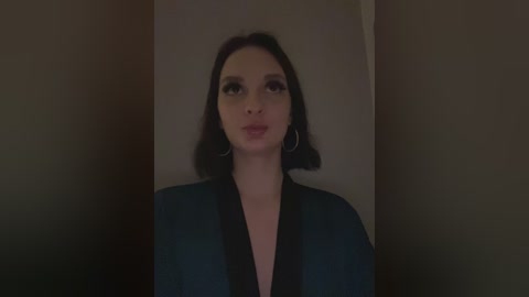 Media: A video of a young woman with straight, dark hair, fair skin, and large hoop earrings, wearing a dark, V-neck top, standing against a dimly lit, plain background.