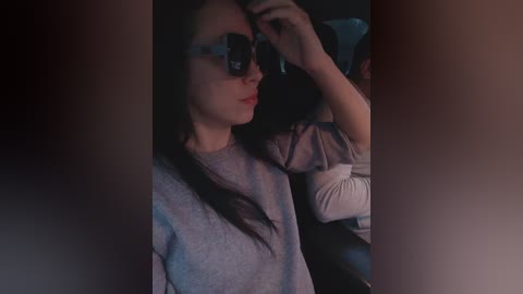 Media: A video of a young woman with long dark hair, wearing sunglasses, a gray sweater, and seated in a dimly lit car, adjusting her glasses.