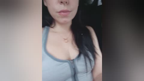 Video of a light-skinned woman with long black hair, wearing a blue tank top, showing cleavage, with a pendant necklace. Background is blurred.