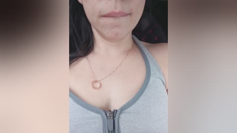 Video of a light-skinned woman with long dark hair, wearing a grey tank top with a zipper, a delicate gold necklace, and a subtle, closed-lip smile.