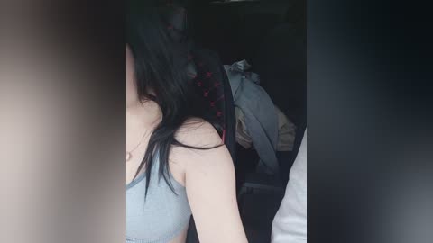 Video of a young woman with long, black hair, wearing a gray tank top, seen from behind, partially obscured by a dark curtain. Background includes a messy room with clothes and a backpack.