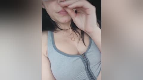 A close-up video of a light-skinned woman with long dark hair, wearing a tight, sleeveless, light blue tank top that accentuates her ample cleavage, partially covering her mouth with her hand.