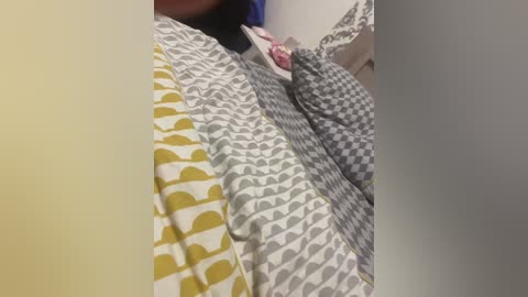 Video of a person's neck and shoulders covered in a patterned, multi-colored duvet. The duvet features yellow, white, and grey geometric shapes. The background is plain and neutral.