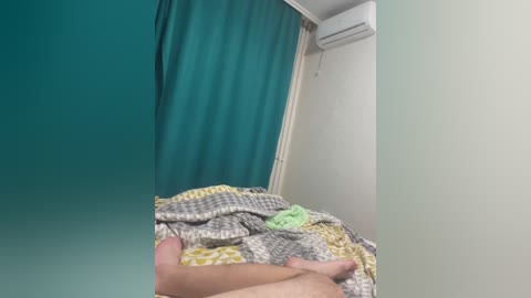 A video of a messy bed with a turquoise curtain, a yellow blanket, and a green pillow in a small, simple room with white walls and an air conditioner.