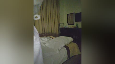 Media: Video of a dimly-lit, sparsely furnished hotel room with a bed covered in white sheets, a beige curtain, and a dark wooden desk with a framed mirror and a green bottle.