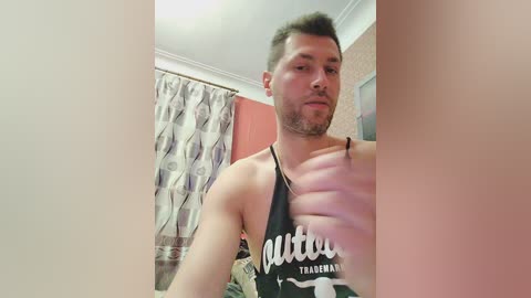 Media: A video of a fit, light-skinned man with short dark hair, wearing a black tank top with white text, standing in a bathroom with beige curtains and pink walls.