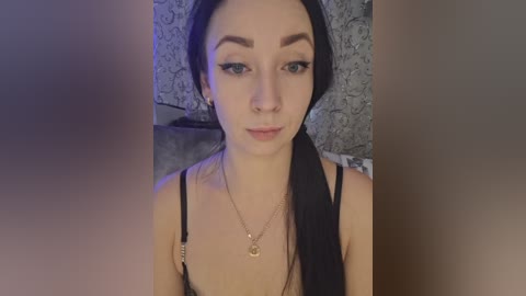 Video of a young woman with pale skin, long black hair, and dark eyebrows, wearing black lace lingerie with thin straps, sitting on a gray patterned couch, holding a selfie stick.