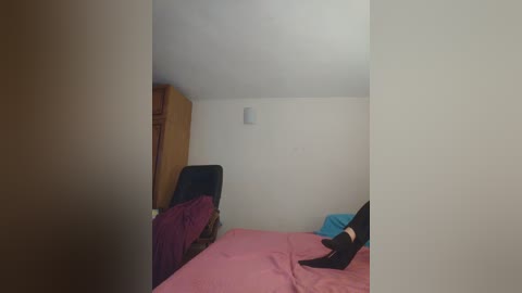 Video of a small, sparsely furnished bedroom with a pink bed, black chair, and wooden closet, featuring a person's legs in black socks and red shoes.