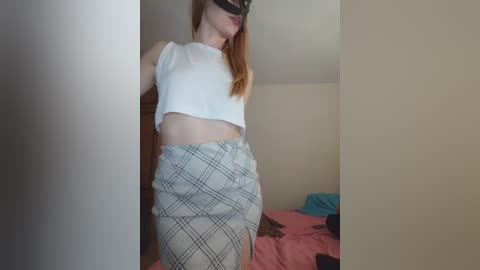 A video of a slender, fair-skinned woman with red hair, wearing a black blindfold and white crop top, standing in a dimly lit bedroom with a pink bedspread.