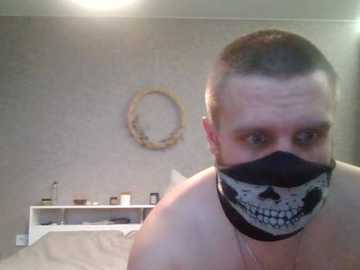 A video of a shirtless man with short hair and a black face mask featuring a white skull design. He stands in a minimalist bedroom with a white bed, shelves, and a circular mirror on the beige wall.