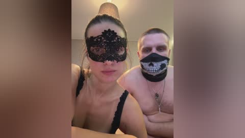 Video of a topless man with a skull face mask and a woman in a black lace mask, both with fair skin, standing closely indoors.