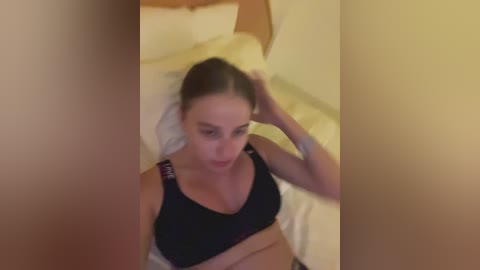 A blurred video of a woman with light skin and long brown hair, wearing a black sports bra and white shirt, sitting on a bed, holding her hair.