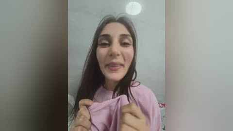 Media: Video of a young woman with long dark hair, fair skin, and a slight smile, holding a light pink shirt against her chest. Background is a plain, light-colored wall.