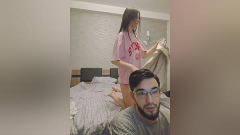 Media: A video shows a man with glasses and a beard in a grey sweater, kneeling on a bed. A woman with long black hair in a pink shirt is standing nearby, holding a blanket.