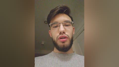 Video of a bearded young man with glasses, wearing a striped shirt, indoors with blurred background.