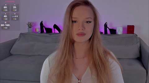 Video of a young Caucasian woman with long blonde hair, wearing a white top, seated on a gray couch. Background includes purple lighting, black high heels, and a digital timer display.