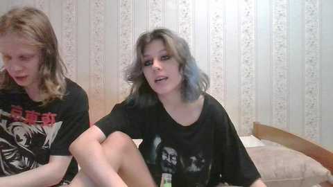 Media: Video of two young women with long hair, one blonde and one blue, wearing black graphic T-shirts, sitting on a bed in a room with striped wallpaper.