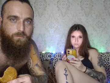 Video of a tattooed, bearded man holding a banana, and a tattooed woman with long hair, both sitting on a leopard-print couch, in a room with floral wallpaper.