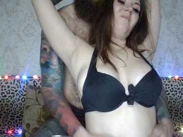 A video of a curvy, fair-skinned woman with long brown hair, wearing a black bra, captured in an intimate embrace with a tattooed man in a leopard print bedspread, surrounded by multicolored string lights.