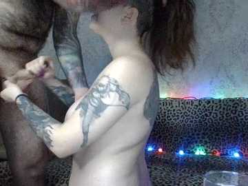 Video of a nude, fair-skinned woman with tattoos, brown hair in a ponytail, and medium-sized breasts, performing oral sex on a dark-skinned man with tattoos on his arm. Background features a leopard-print couch with multicolored fairy lights.
