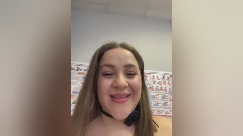 A video of a young woman with light skin, long brown hair, and a black choker, smiling, standing in front of a wall with colorful anatomical posters.