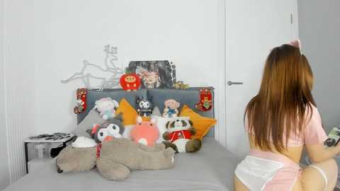 Media: Video of a young woman with long brown hair in pink pajamas facing a cluttered toy shelf with plush toys, including a panda and a red bear, in a minimalist bedroom.