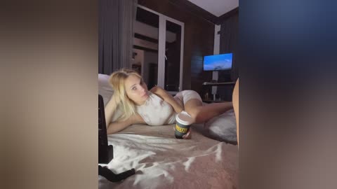 Media: Video of a blonde woman in a white tank top and shorts lying on a bed, holding a black remote, with a coffee cup beside her.