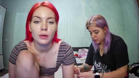 Media: Video of two young women: a red-haired woman with piercings, and a blonde woman with purple hair, in a bedroom with pale green wallpaper.