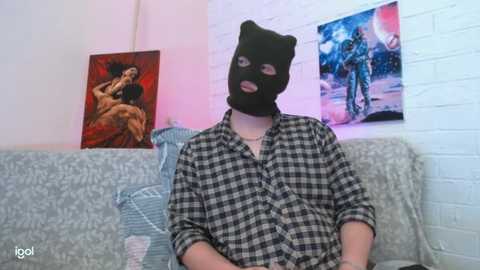 Video of a person wearing a black balaclava and plaid shirt sitting on a couch in a room with red and blue artwork on the white brick wall.