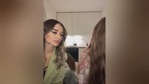 Media: Video of a woman with long, wavy, light brown hair and fair skin, wearing a green jacket, standing in a modern kitchen with white cabinets and a tiled backsplash.