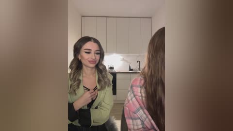 Media: Video of two women: a brunette with wavy hair in a green jacket, holding a black bag, and a blonde in a pink plaid shirt, standing in a modern kitchen with white cabinets and a sink.
