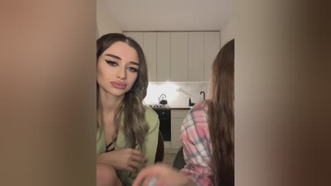 Media: Video of a young woman with long, wavy brown hair, wearing a green blouse, sitting in a modern kitchen with white cabinets, a sink, and a stove. Another person, partially visible, is wearing a plaid shirt.