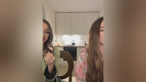 Media: Video of two young women with long brown hair, seated at a table in a modern kitchen. One woman, in a green coat, holds a phone, while the other wears a pink plaid shirt.