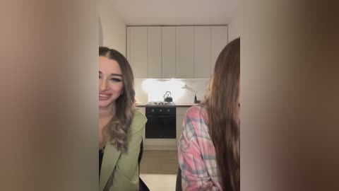 Media: Video of two young women in a modern kitchen. One with long, wavy blonde hair, green blazer, smiling, standing. The other, with long, straight brown hair, wearing a plaid shirt, seated at a counter.