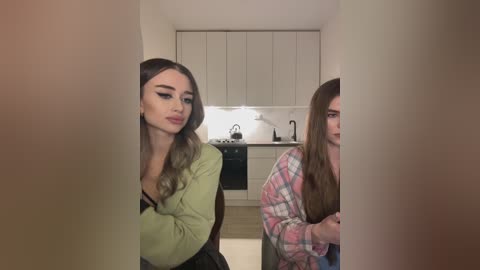 Media: Video of two women in a kitchen, one with long, wavy brown hair in a green top, the other with long, straight brown hair in a plaid shirt, looking away.