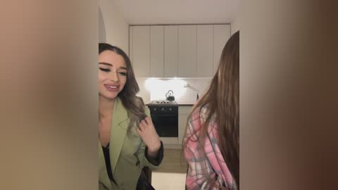 Media: Video of two women in a modern kitchen: One, with long brown hair, wears a green blazer, grinning; the other, in a plaid shirt, has her back to the camera.