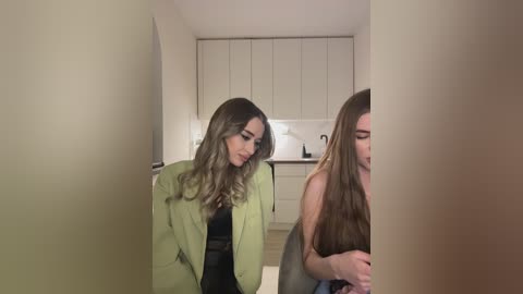 Media: Video of two women in a modern kitchen. One wears a light green blazer, black top, and long hair, the other has long brown hair and a gray top.