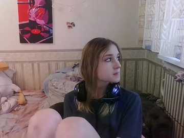 Media: Video of a young woman with light skin and shoulder-length brown hair, wearing headphones, sitting on a bed in a cluttered room with a colorful painting and a window with a white curtain.