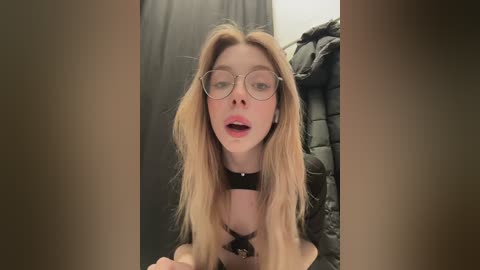 A video of a fair-skinned, blonde woman with glasses and a black choker, sitting in a dimly lit room with black curtains and a pile of clothes in the background.
